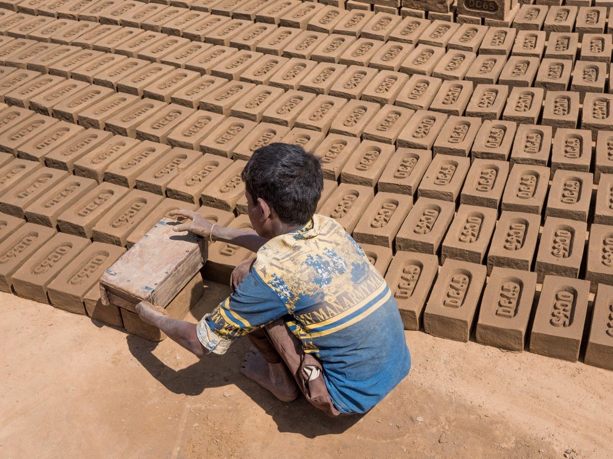 Manuelle Ziegelproduktion Eine Person legt einen Lehmziegel aus einer Form neben Reihen trocknender Ziegel auf staubigem Boden ab.