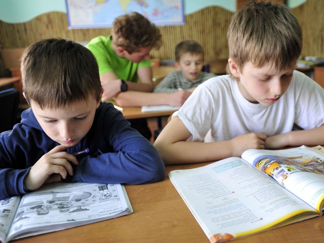 Vier Jungen im Klassenzimmer, zwei im Vordergrund konzentrieren sich intensiv auf ihre offenen Schulbücher.