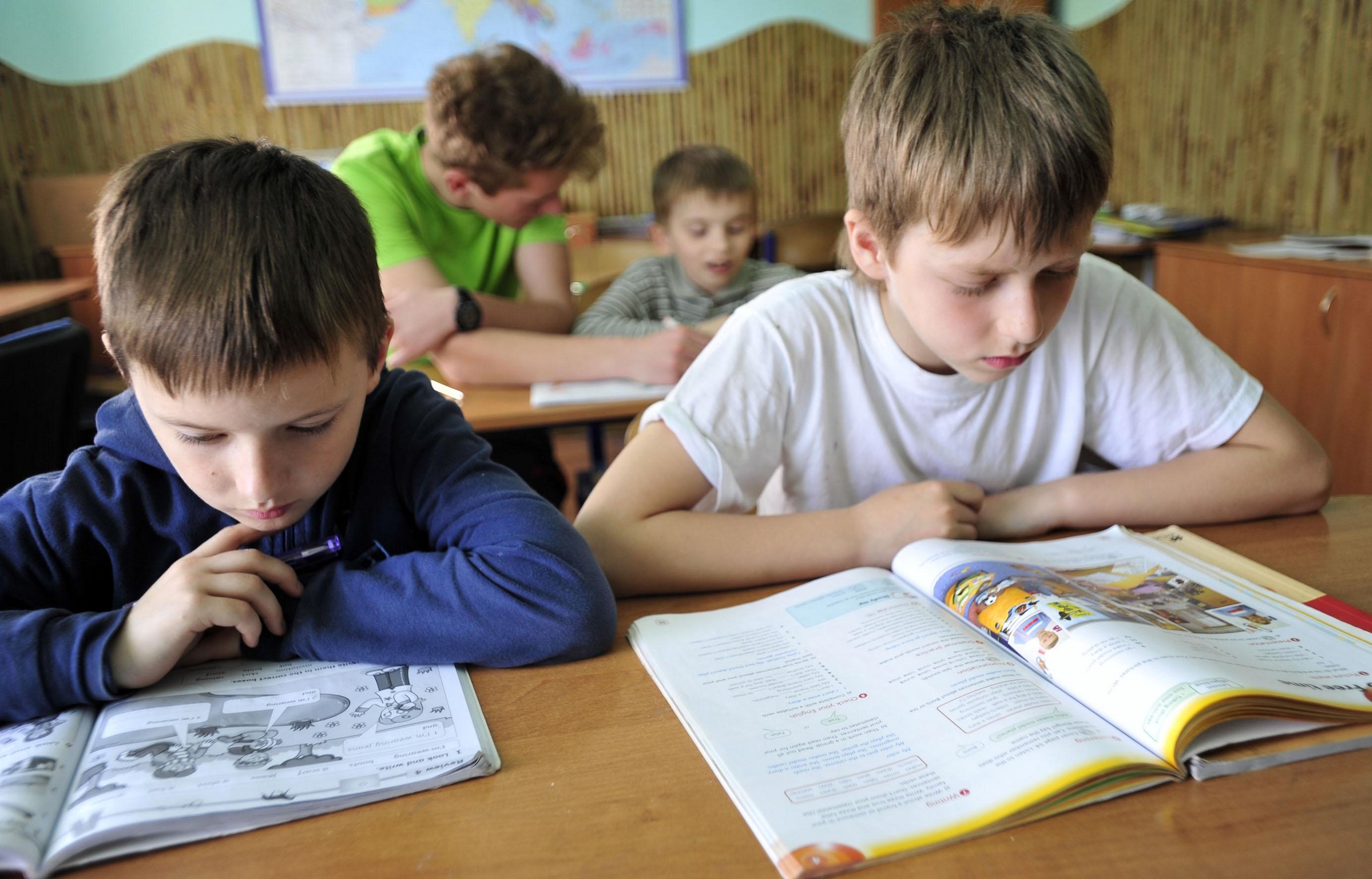 Vier Jungen im Klassenzimmer, zwei im Vordergrund konzentrieren sich intensiv auf ihre offenen Schulbücher.