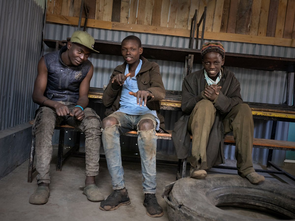 Drei lächelnde junge Männer sitzen auf einer Bank in einem einfachen Raum mit Metallwänden, einer zeigt nach vorne.