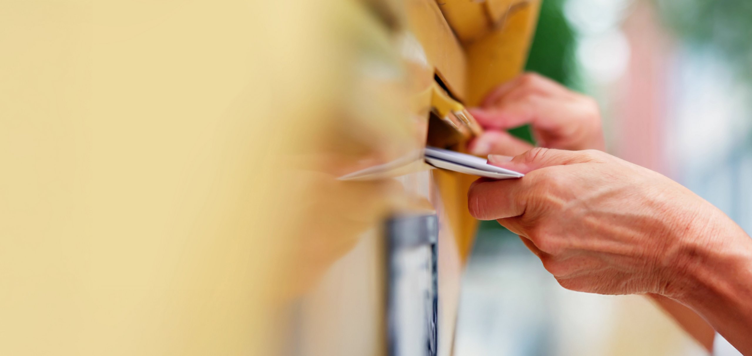 Nahaufnahme einer Hand, die Post, möglicherweise Briefe oder ein kleines Paket, in einen gelben Briefkasten steckt.