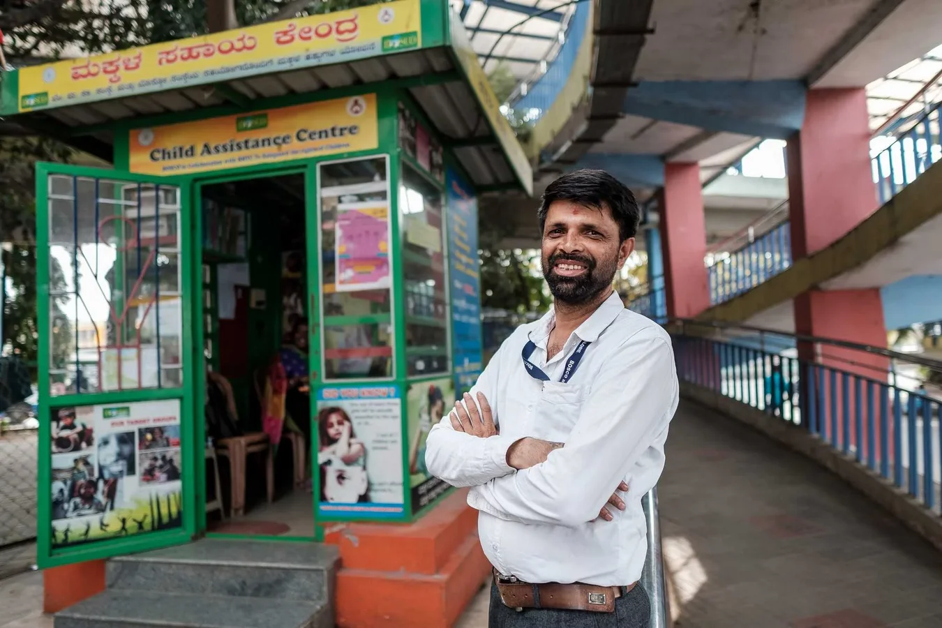 Lächelnder bärtiger Mann in weißem Hemd mit verschränkten Armen vor einem grünen Stand des "Kinderhilfezentrums".