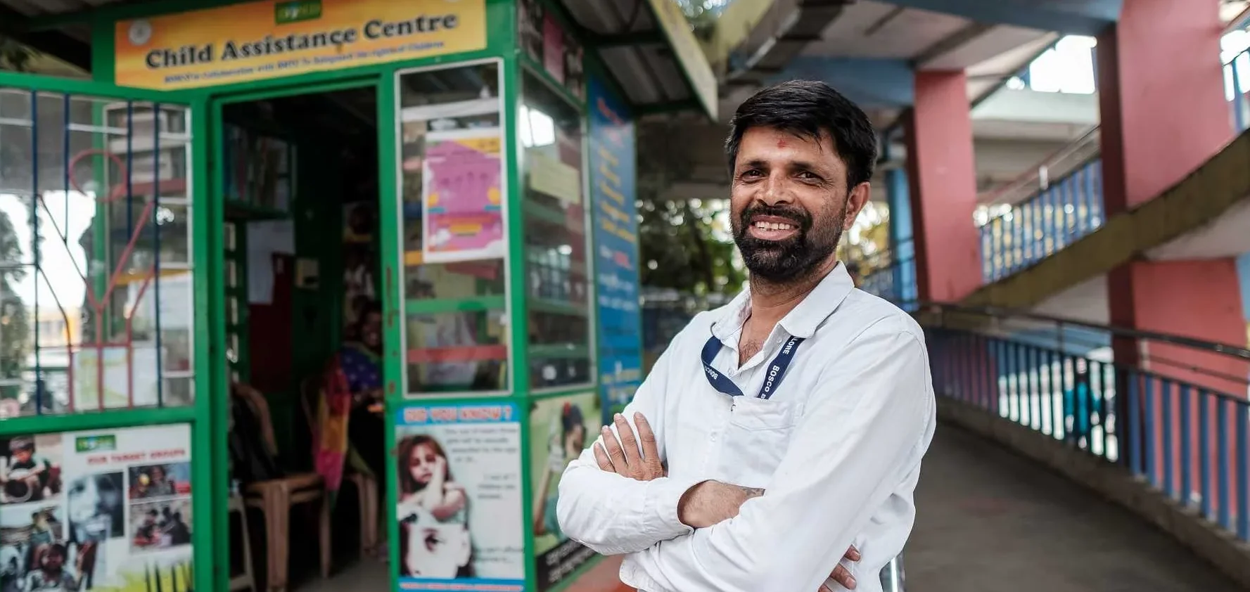Lächelnder bärtiger Mann in weißem Hemd mit verschränkten Armen vor einem grünen Stand des "Kinderhilfezentrums".