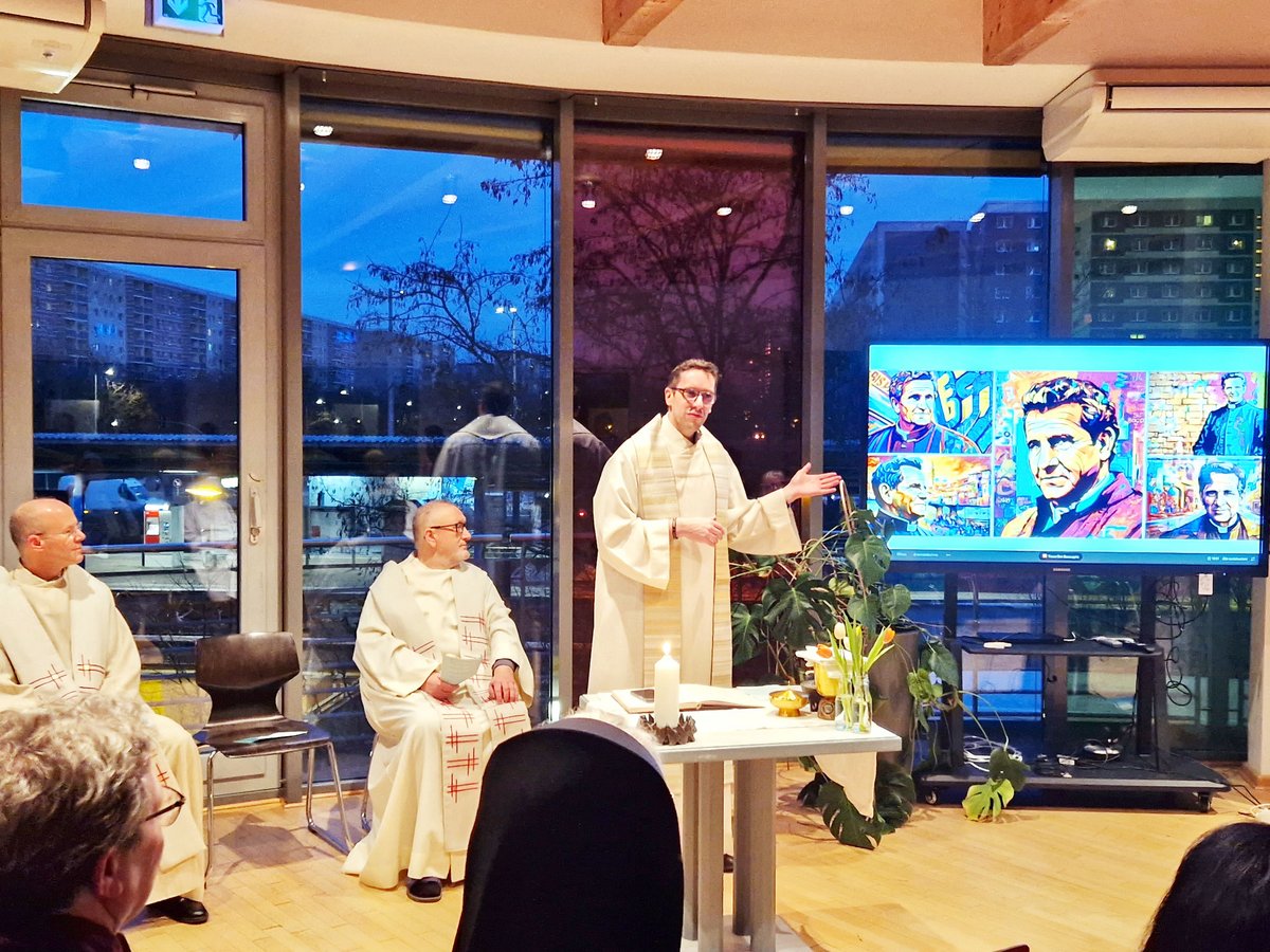 Ein Priester in weißen Gewändern präsentiert vor einer Gemeinde, mit anderen Klerikern sitzend, in einem Raum mit großen Fenstern.