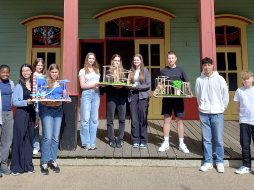 Eine Gruppe Schüler präsentiert ihre selbstgebauten Achterbahn- und Architekturmodelle auf einer Veranda.