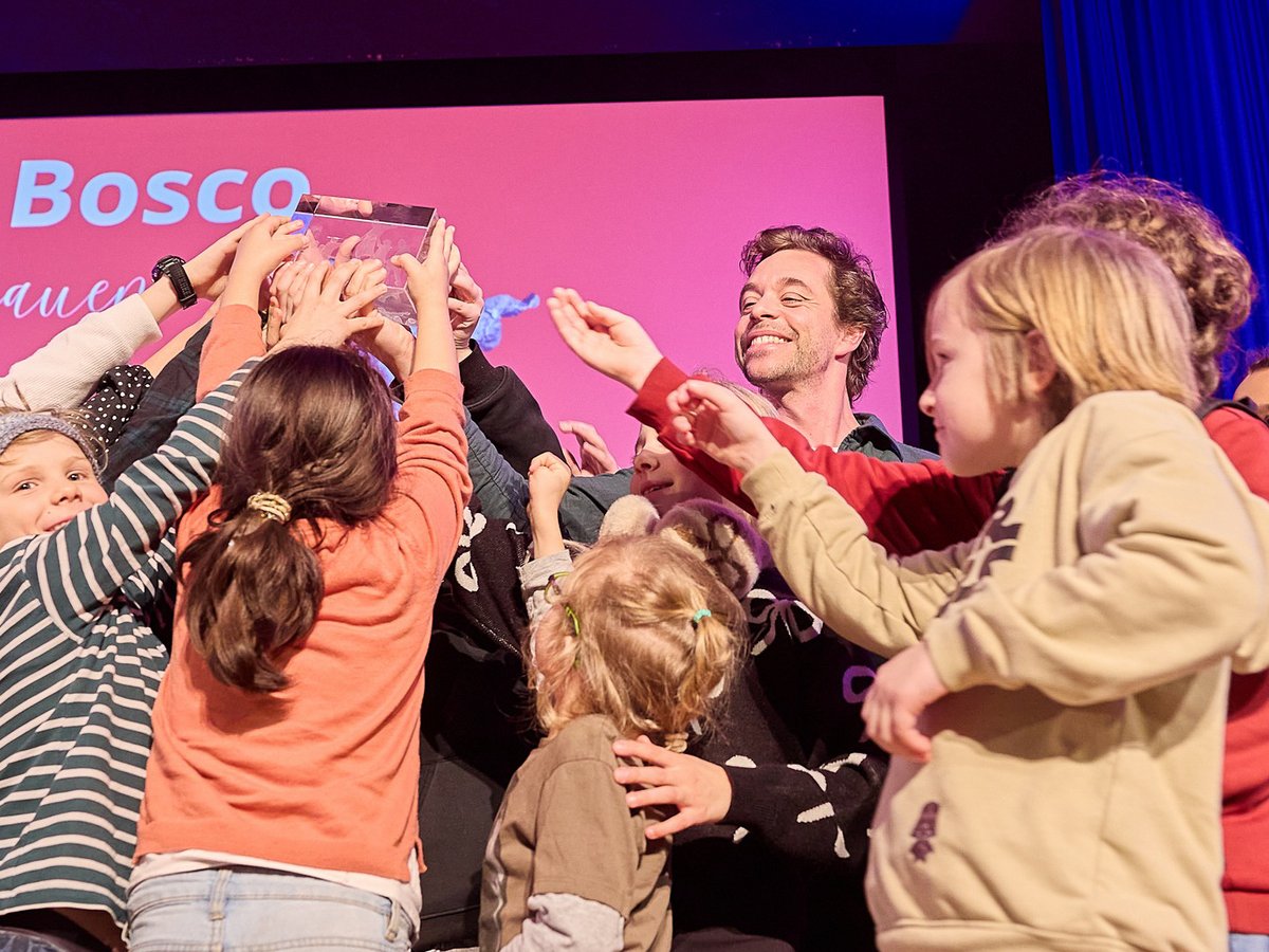 Lächelnder Mann und aufgeregte Kinder auf der Bühne, die gemeinsam eine durchsichtige Trophäe halten und einen Erfolg feiern.