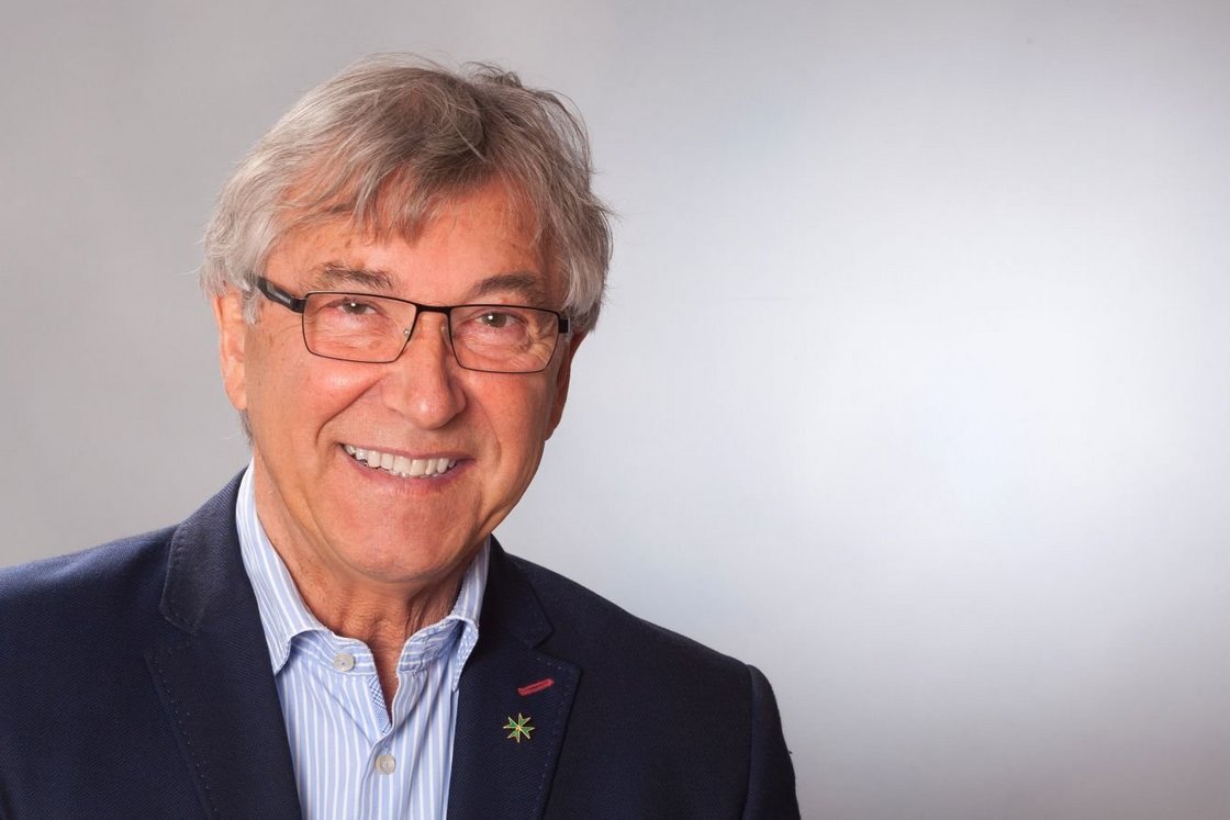 Lächelnder älterer Mann mit grauem Haar und Brille, trägt ein blaues Sakko mit einer kleinen Sternnadel.