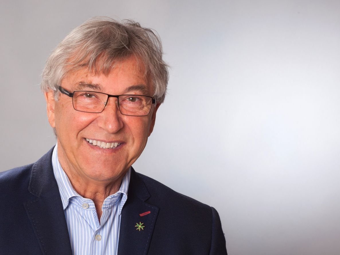 Lächelnder älterer Mann mit grauem Haar und Brille, trägt ein blaues Sakko mit einer kleinen Sternnadel.