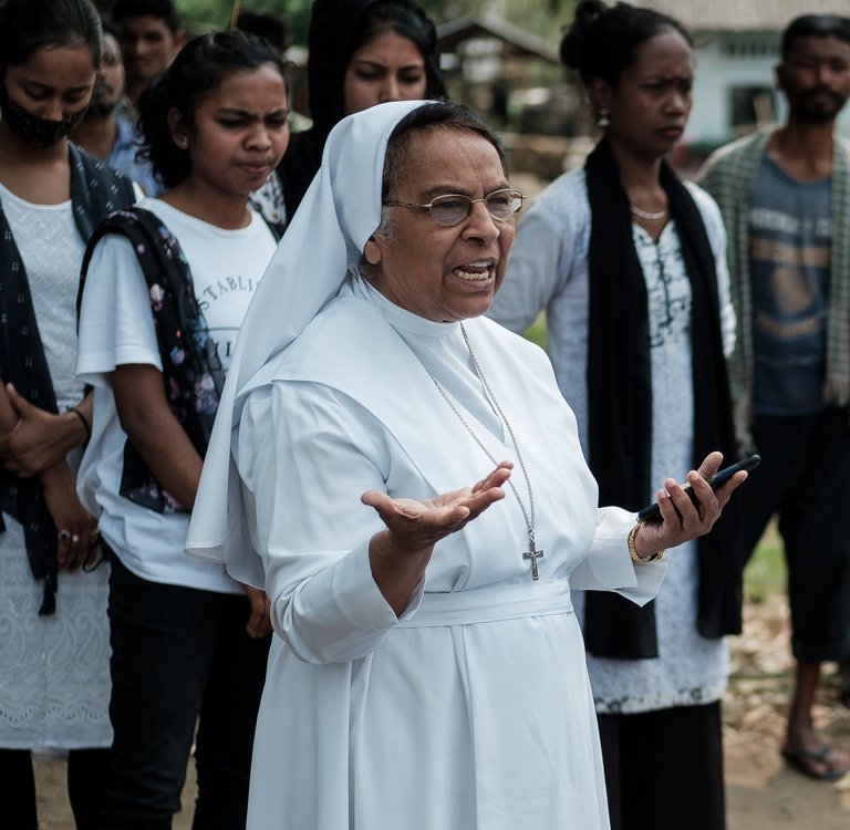 Eine katholische Nonne im weißen Habit spricht zu einer vielfältigen Gruppe von Menschen, darunter Frauen und Kinder, im Freien in einem Dorf.