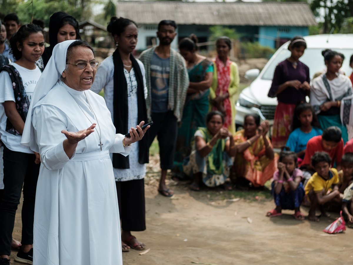 Eine katholische Nonne im weißen Habit spricht zu einer vielfältigen Gruppe von Menschen, darunter Frauen und Kinder, im Freien in einem Dorf.