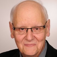 Kopfaufnahme eines lächelnden älteren Mannes mit Brille, weißem Haar, grauem Hemd, gestreifter Krawatte und schwarzem Sakko.