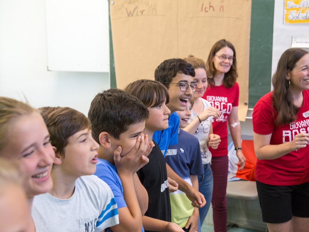 Eine Reihe lachender Kinder und Erwachsener, einige in roten 'Giovanni Bosco'-Shirts, bei einer lebhaften Klassenaktivität.