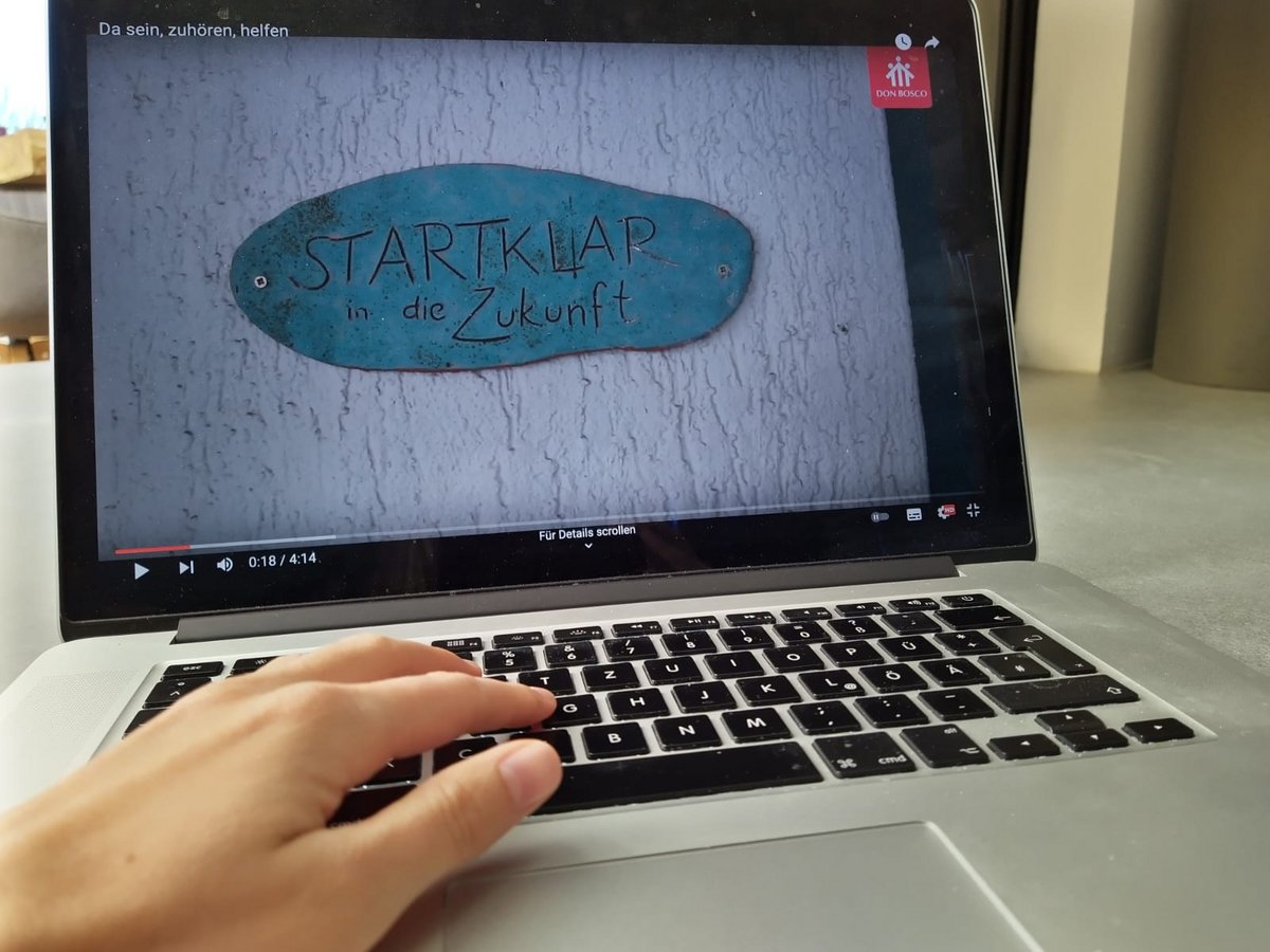 Laptop zeigt Video: 'Da sein, zuhören, helfen', 'STARTKLAR in die Zukunft' und Don Bosco Logo. Hand auf Tastatur.