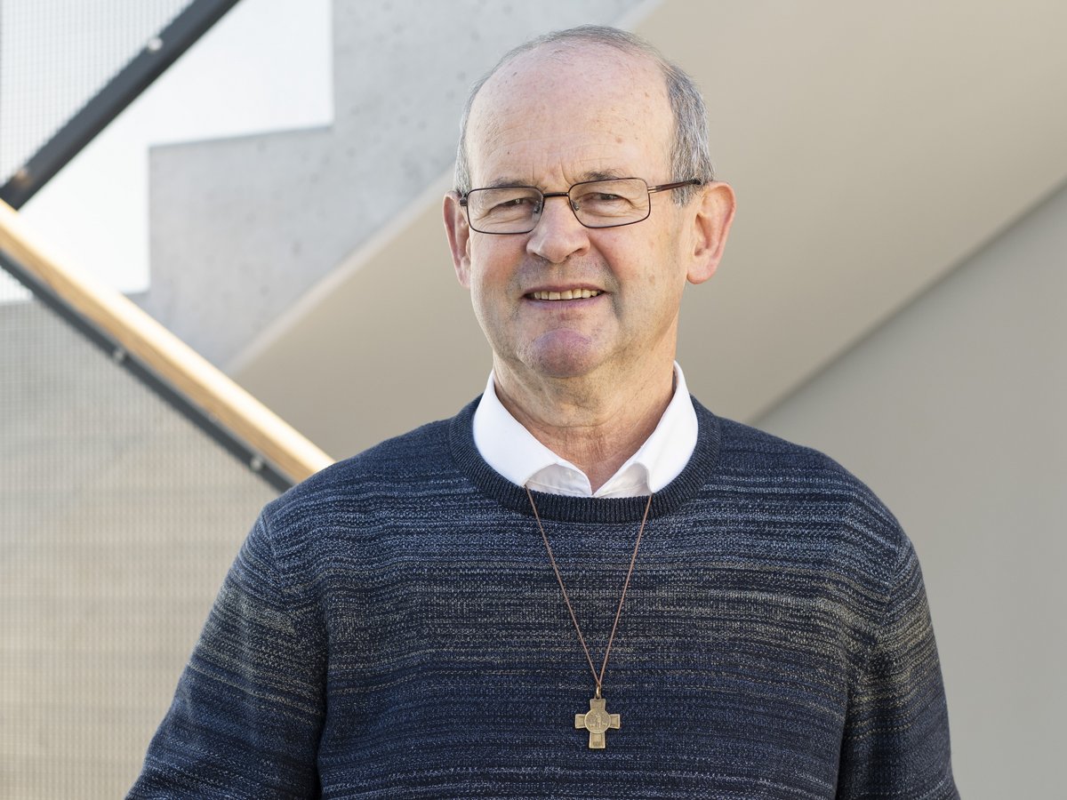 Ein lächelnder älterer Mann mit Brille und Kreuzanhänger steht an einem rot-weißen Geländer vor einer modernen Treppe.
