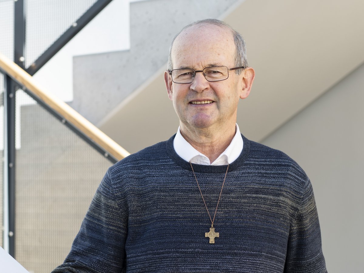 Ein lächelnder älterer Mann mit Brille und Kreuzanhänger steht an einem rot-weißen Geländer vor einer modernen Treppe.