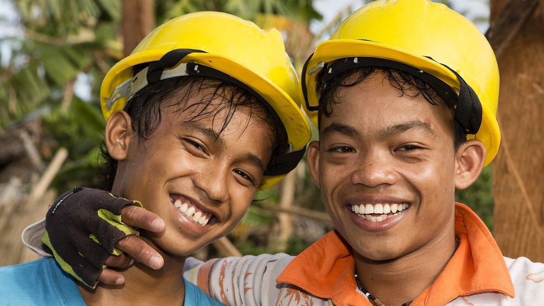 Zwei lächelnde junge Männer mit gelben Schutzhelmen, verschwitzt, einer mit behandschuhter Hand auf der Schulter des anderen.