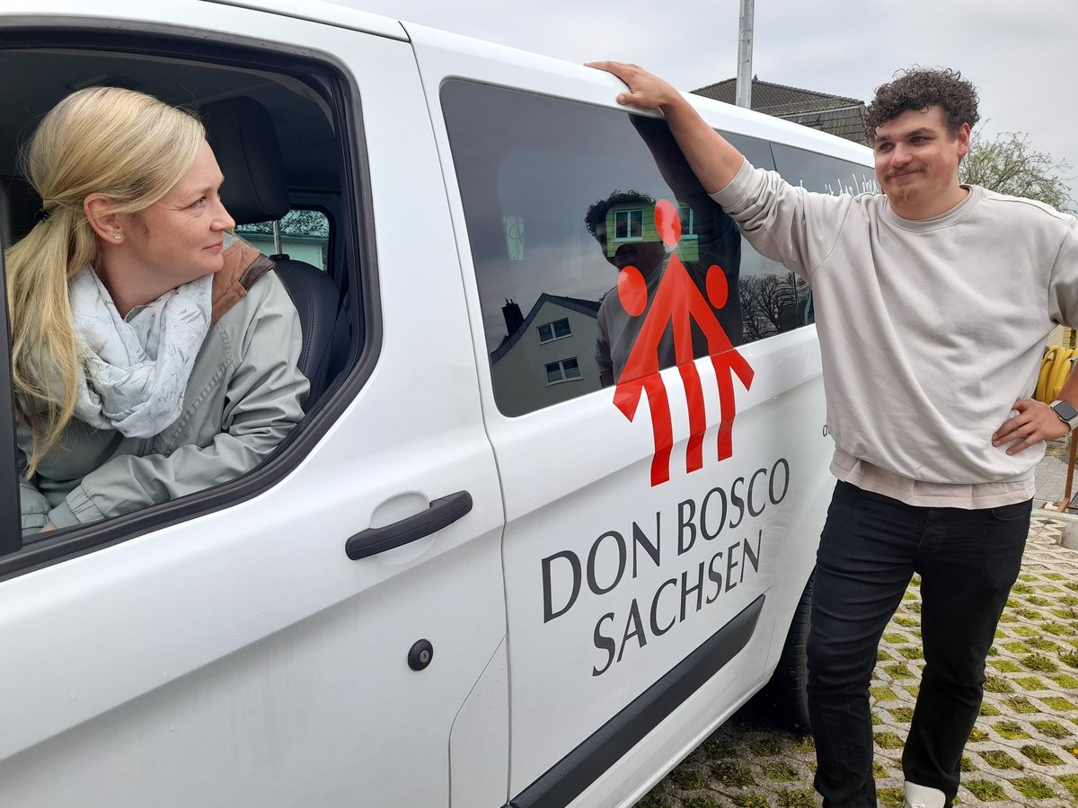 Eine Frau in einem weißen 'Don Bosco Sachsen' Transporter blickt einen Mann mit lockigem Haar an, der sich anlehnt.