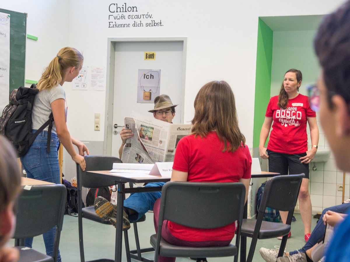Menschen in einem Klassenzimmer, darunter eine Frau im roten Hemd sprechend, ein Mann Zeitung lesend, andere sitzend.