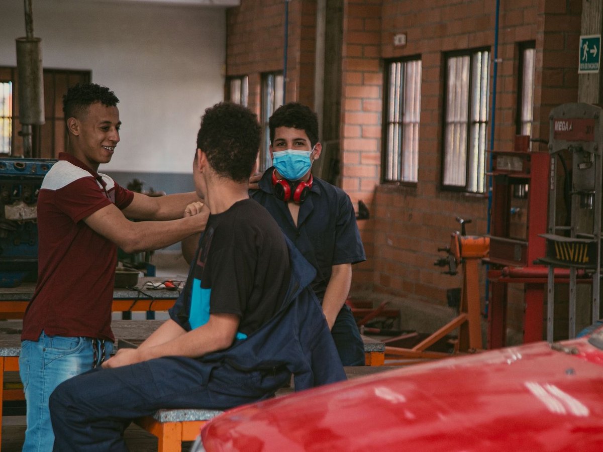 Drei junge Männer in einer Werkstatt: einer lächelt, einer sitzt, und einer mit Maske und Kopfhörern blickt zur Kamera.