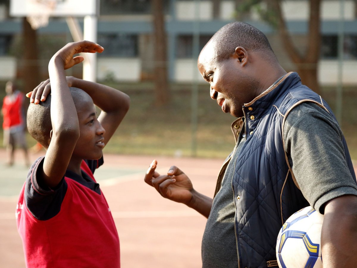 Ein erwachsener Trainer mit Fußball spricht mit einem jungen Jungen auf einem Sportplatz; der Junge hört aufmerksam zu.