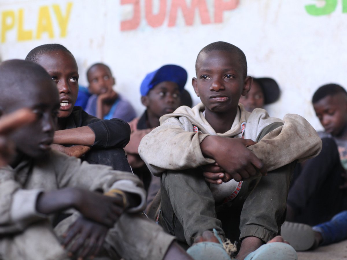Gruppe junger afrikanischer Jungen sitzt auf dem Boden, einige aufmerksam blickend, mit 'PLAY JUMP SING' an der Wand dahinter.