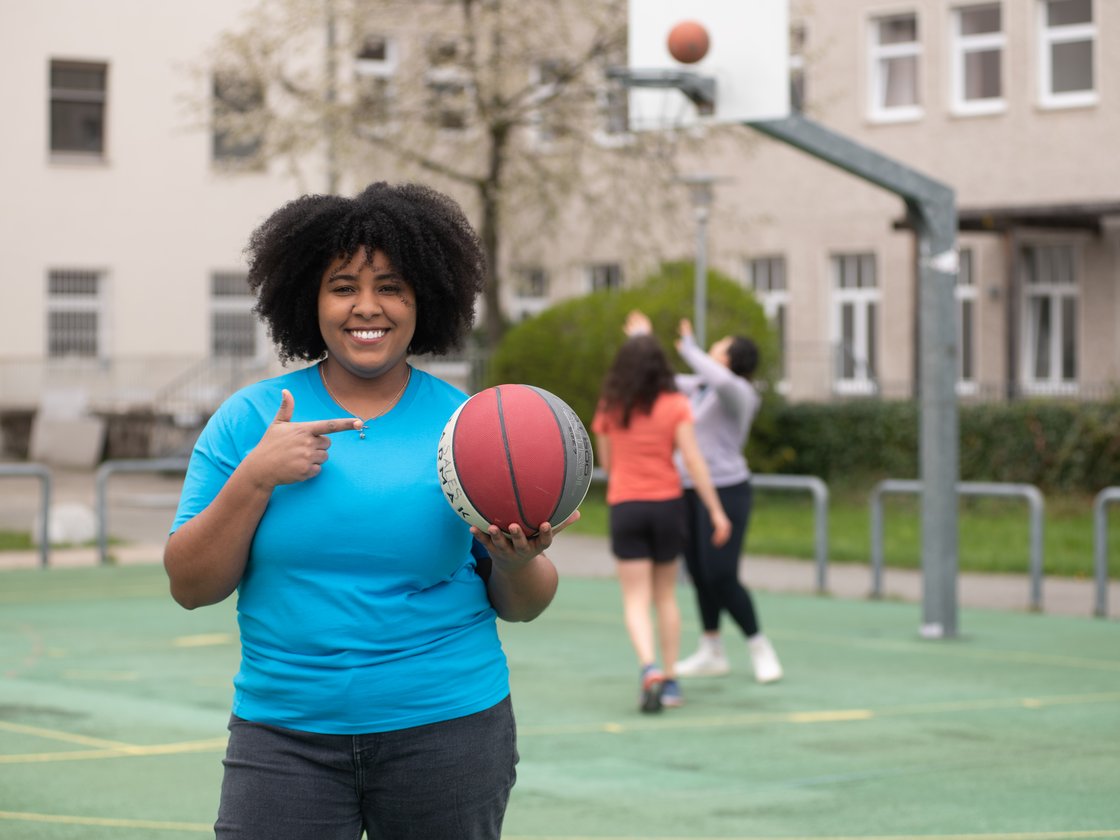 Lächelnde Schwarze Frau mit lockigem Haar im blauen T-Shirt hält Basketball und zeigt darauf, andere spielen im Hintergrund.