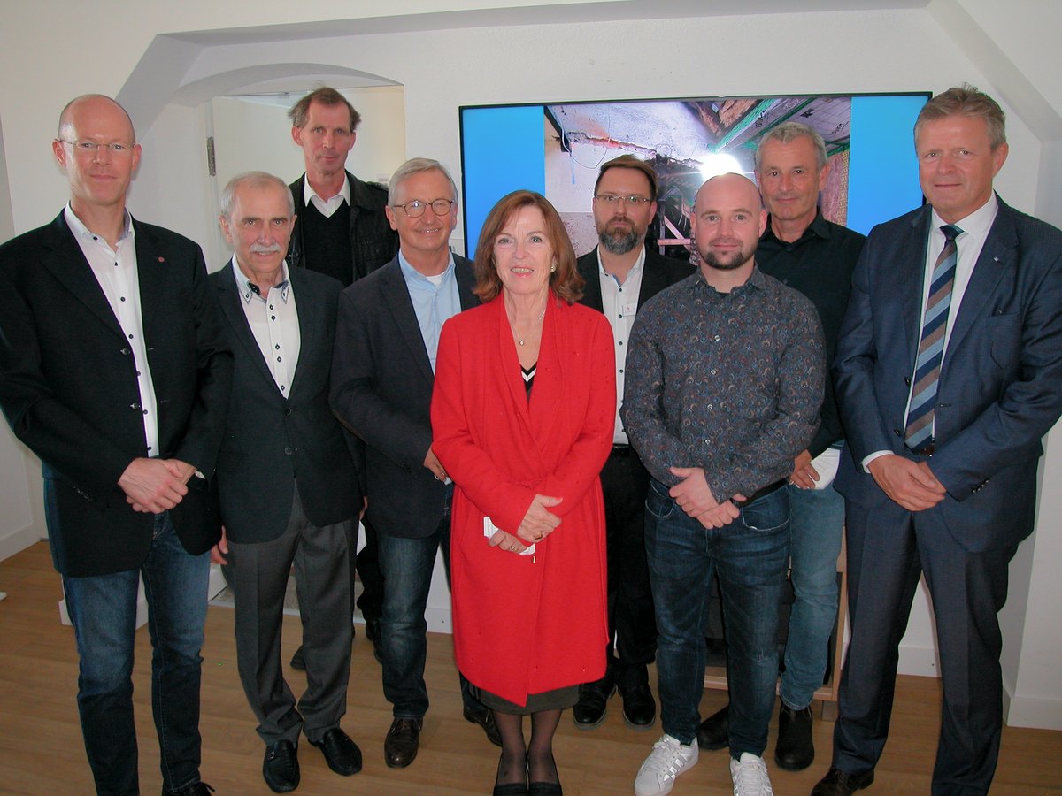 Eine Gruppe von zehn Personen, neun Männer und eine Frau im roten Mantel, posieren drinnen für ein Foto, mit einem Bildschirm im Hintergrund.