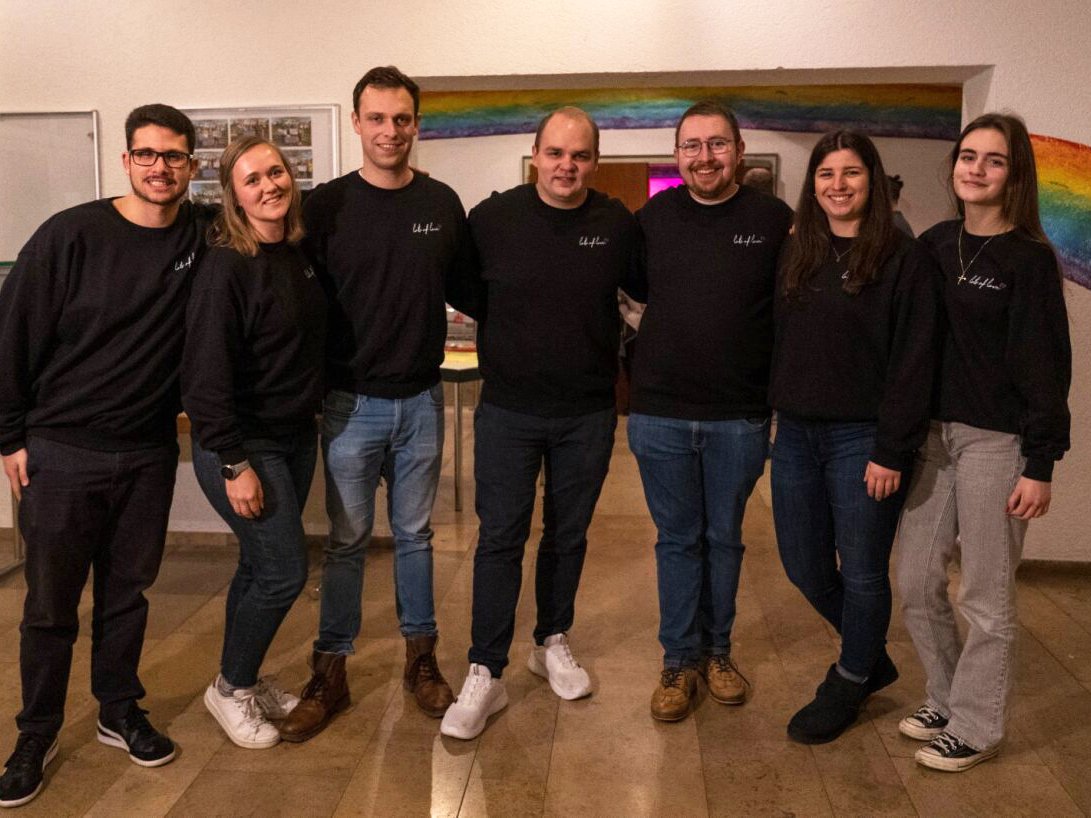 Sieben lächelnde Personen in schwarzen Sweatshirts und Jeans posieren vor einer Wand mit einem Regenbogen-Wandgemälde.