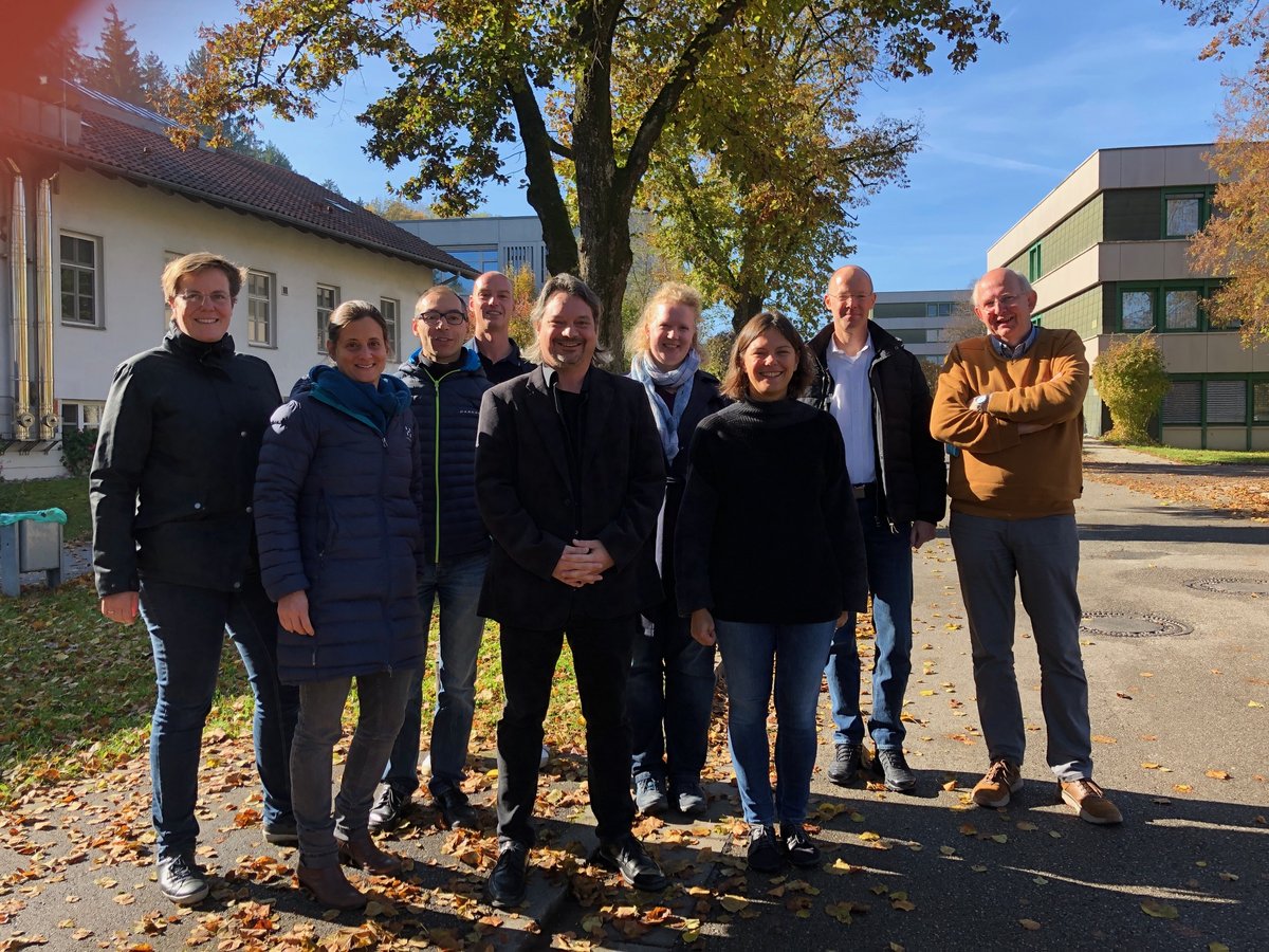 Eine Gruppe von zehn lächelnden Personen steht an einem sonnigen Herbsttag im Freien vor Gebäuden und Bäumen mit Herbstlaub.