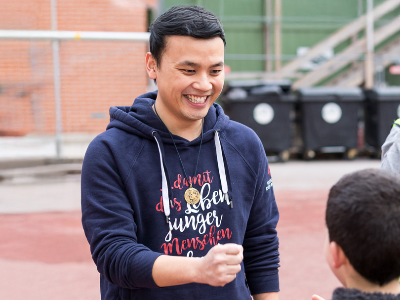Lächelnder Mann im blauen Hoodie mit „junger menschen“-Aufschrift und Medaillon, spielt mit Kind auf rotem Platz.
