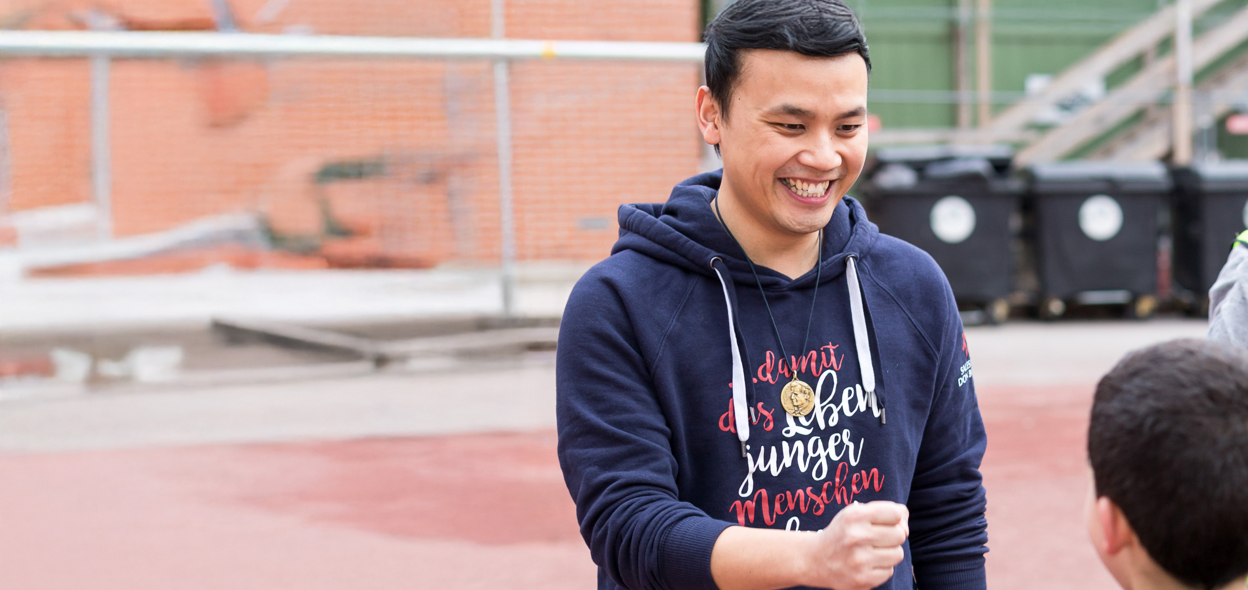 Lächelnder Mann im blauen Hoodie mit „junger menschen“-Aufschrift und Medaillon, spielt mit Kind auf rotem Platz.