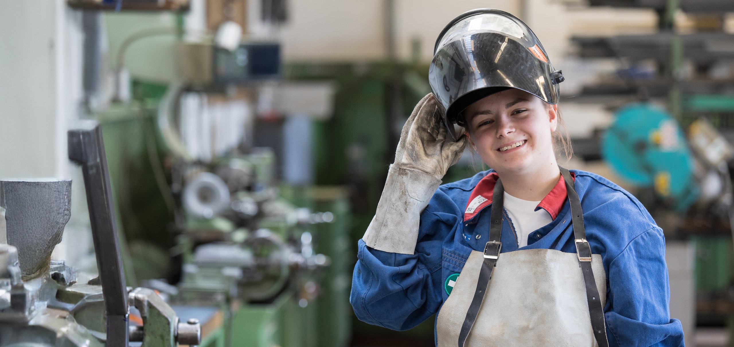 Lächelnde junge Frau in Schweißerausrüstung mit hochgeschobenem Helm, in einer Werkstatt mit Maschinen im Hintergrund.