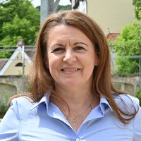 Porträt einer lächelnden Frau mit braunen Haaren und hellblauem Hemd, die eine Kreuzkette trägt, vor verschwommenem grünem Hintergrund.