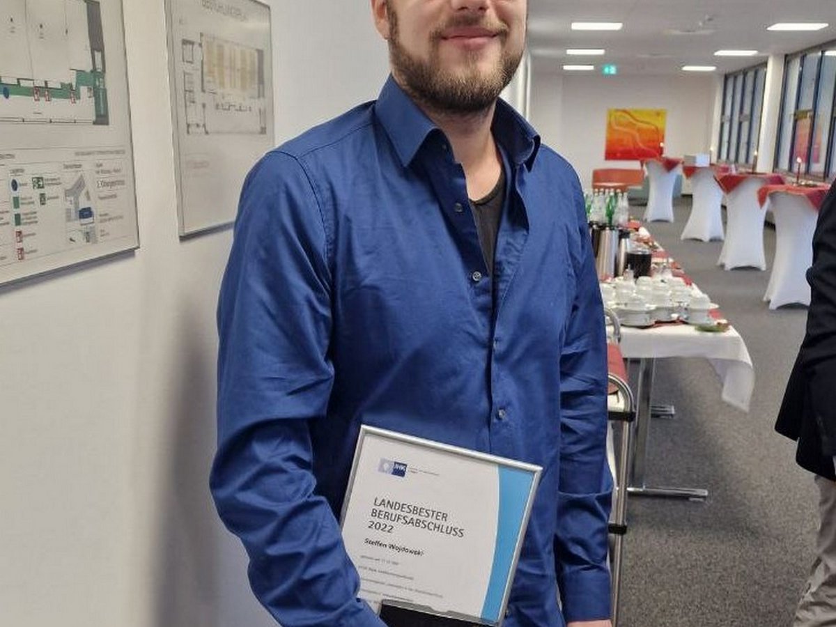 Steffen Wojdowski, ein junger Mann mit Bart und Brille, lächelt und hält seine Urkunde als Landesbester Berufsabschluss 2022.