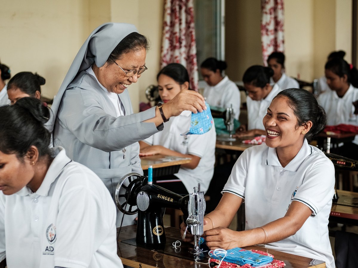 Eine lachende Nonne zeigt einer lächelnden Schülerin Stoff an einer Nähmaschine. Andere Frauen nähen auch.