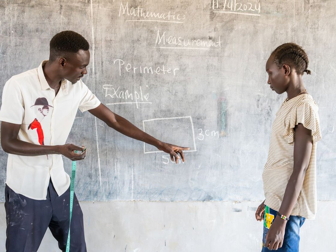 Mathelehrer zeigt auf ein Rechteck an einer Tafel, erklärt den Umfang mit einem Maßband einer Schülerin.