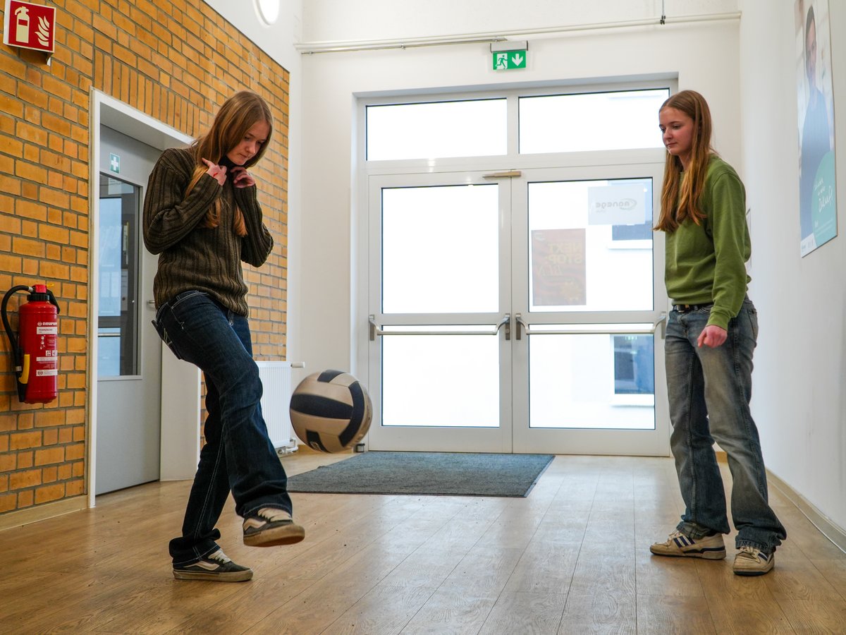 Zwei junge Frauen in einem Flur. Eine kickt einen Volleyball, die andere schaut zu. Backsteinwand und Glastüren im Hintergrund.
