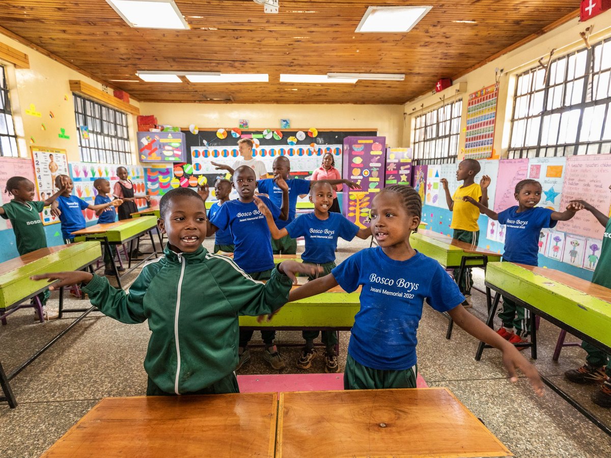 Junge Schüler und ein Erwachsener strecken im farbenfrohen Klassenzimmer die Arme aus, viele tragen „Bosco Boys“-Shirts.