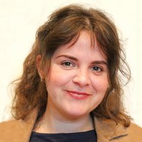 Eine lächelnde Frau mit lockigem braunem Haar trägt ein dunkles Oberteil und eine hellbraune Jacke und blickt den Betrachter an.