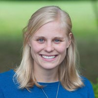 Eine blonde Frau mit blauen Augen lächelt, trägt ein blaues Oberteil und eine kleine Halskette vor grünem Hintergrund.