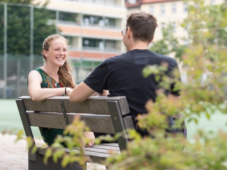 Eine junge Frau mit fröhlichem Lächeln spricht mit einem Mann auf einer Holzparkbank. Grüne Blätter im Vordergrund.