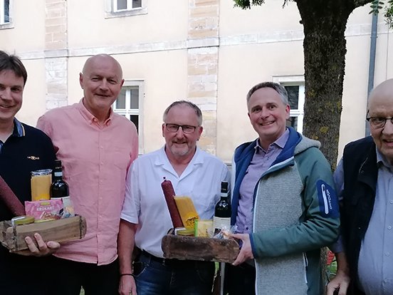 Fünf lächelnde Männer halten Geschenkkörbe mit Würsten, Flaschen und Lebensmitteln vor einem Gebäude.