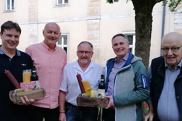 Fünf lächelnde Männer halten Geschenkkörbe mit Würsten, Flaschen und Lebensmitteln vor einem Gebäude.