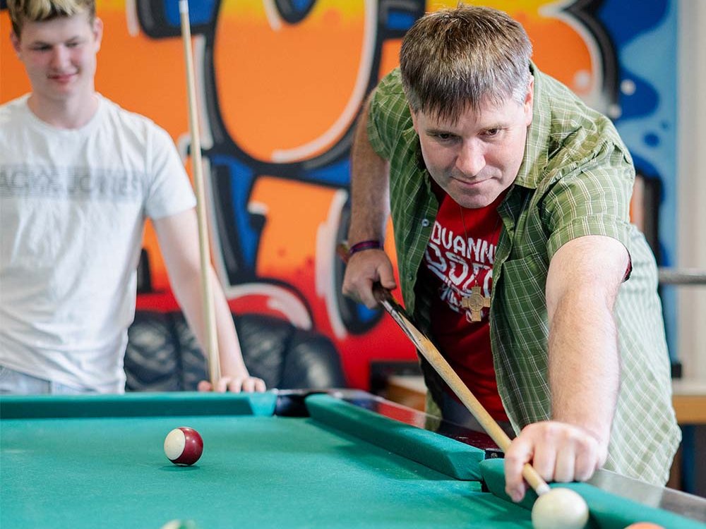 Ein Mann im grünen Hemd spielt Billard und zielt auf die weiße Kugel. Ein jüngerer Mann schaut im Hintergrund zu, vor einer Graffitiwand.