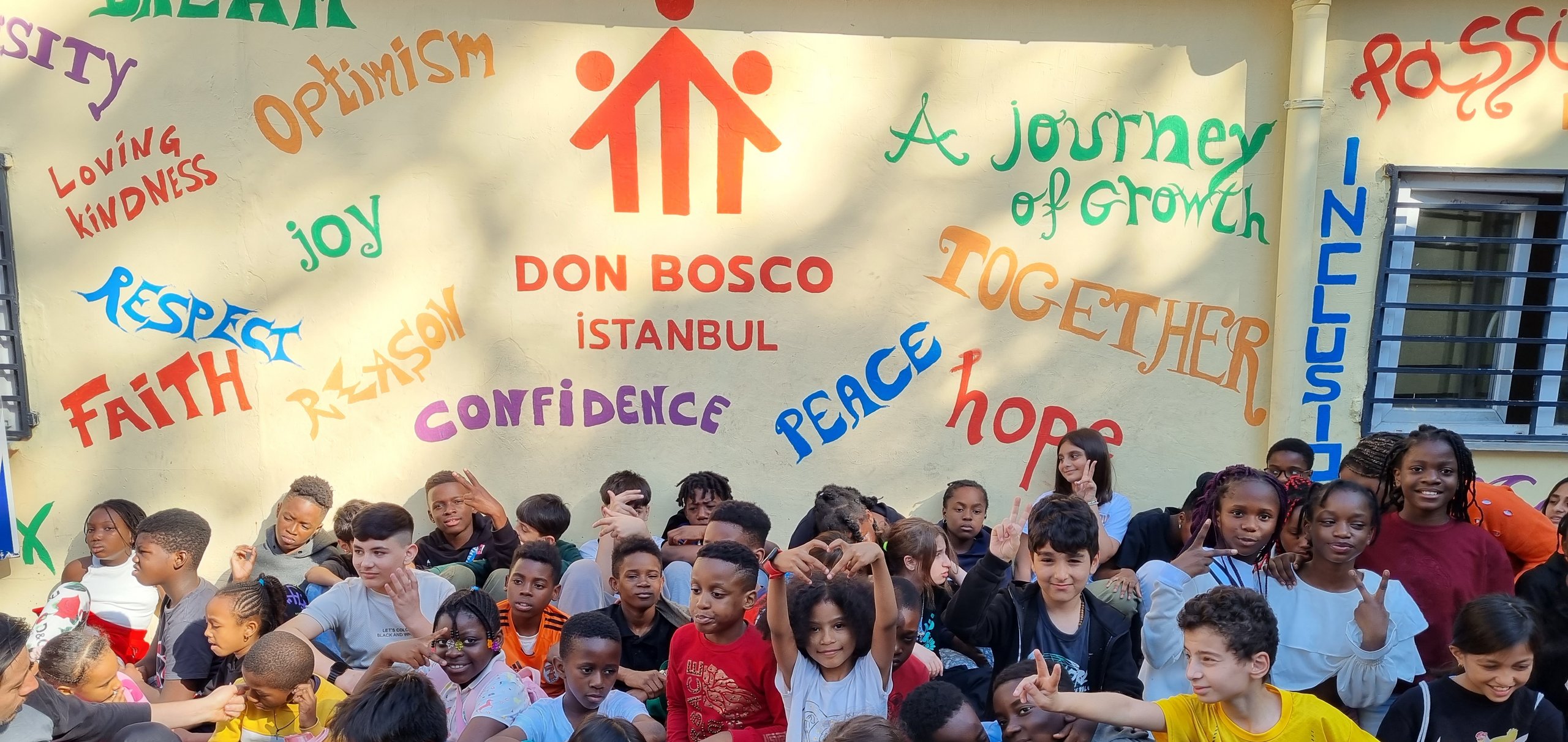 Vielfältige Gruppe von Kindern und Jugendlichen sitzt vor einem bunten Wandgemälde mit Wörtern wie 'Traum', 'Optimismus', 'Frieden' und 'Hoffnung'.