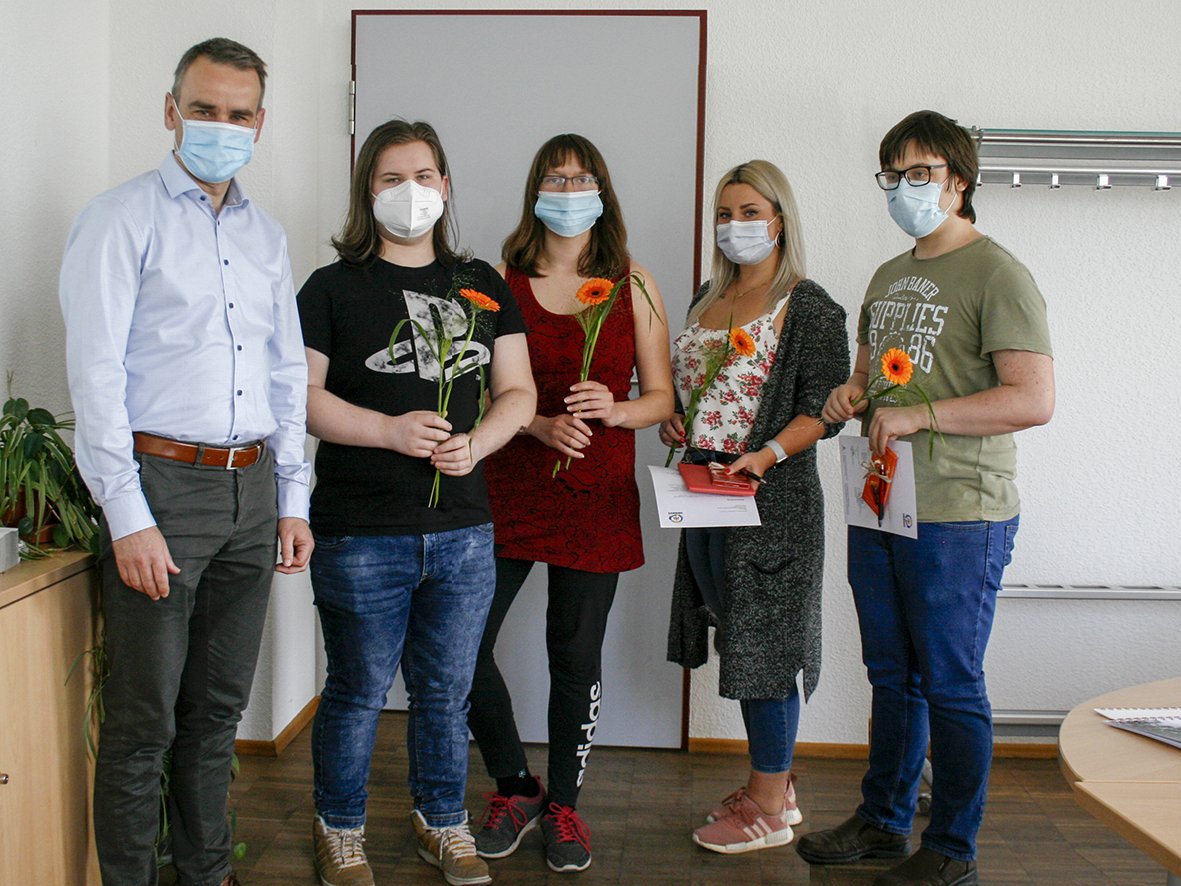 Fünf maskierte Personen, darunter ein Mann und vier jüngere Leute, stehen zusammen und halten orange Gerbera und Zertifikate.