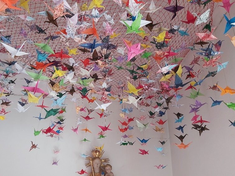 Hunderte bunter Origami-Papierkraniche hängen an einem roten Netz, das eine Deckenverzierung über einer Marienstatue mit Jesuskind bildet.