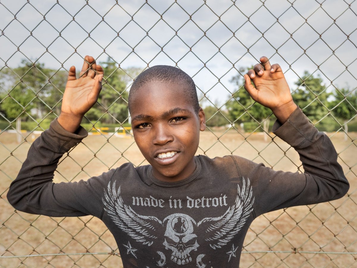 Porträt eines jungen Jungen, der hinter einem Maschendrahtzaun lächelt, ein 'made in Detroit'-Shirt trägt und die Hände hebt.