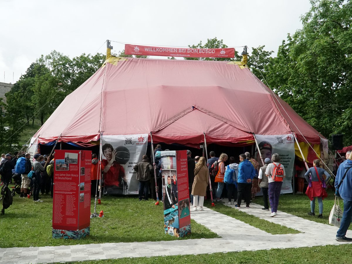 Rotes Festzelt mit einem "Willkommen bei Don Bosco"-Banner auf grünem Rasen, viele Besucher strömen in den Eingangsbereich.