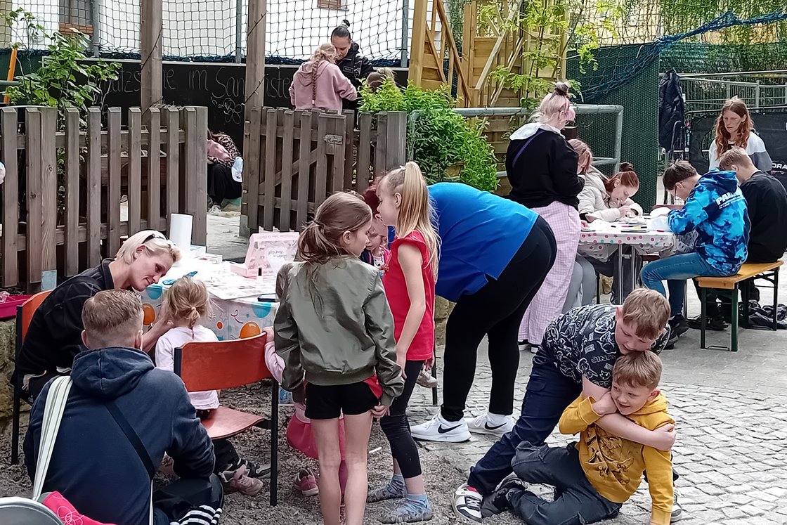 Ein lebhaftes Outdoor-Event mit Erwachsenen und Kindern. Einige basteln an Tischen, andere spielen auf dem Boden.
